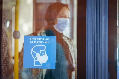 Eine Frau mit einer Mund-Nasen-Bedeckung steht in einem Bus. Ein Schild weist darauf hin, dass im ÖPNV Maskenpflicht gilt.